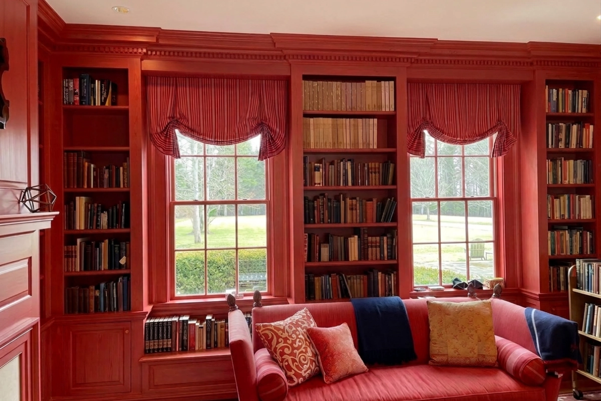 Red-paneled library with built-in bookshelves, large windows, and a red sofa with colorful pillows.